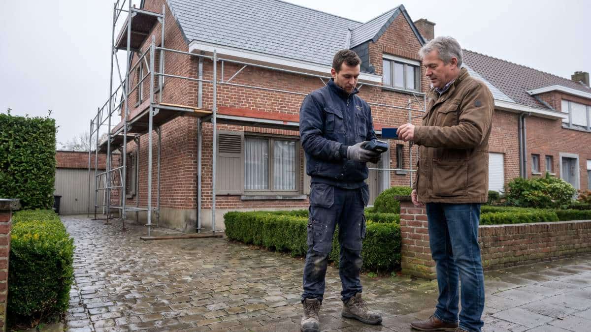 Homme payant avec sa carte sans contact sur un terminal de paiement auprès d'un ouvrier effectuant des travaux sur une maison en Belgique.